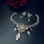 Elegant handcrafted silver necklace set with matching earrings on a dark background, surrounded by flowers.