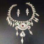 Traditional silver-plated necklace set with a central floral motif, red and green enamel teardrops, and matching drop earrings, displayed on a black background.