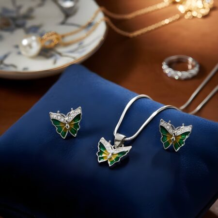 A silver jewelry set featuring a butterfly design with green enamel detailing. The set includes a butterfly-shaped pendant on a silver chain and a matching pair of stud earrings, both accented with sparkling stones on the wings.