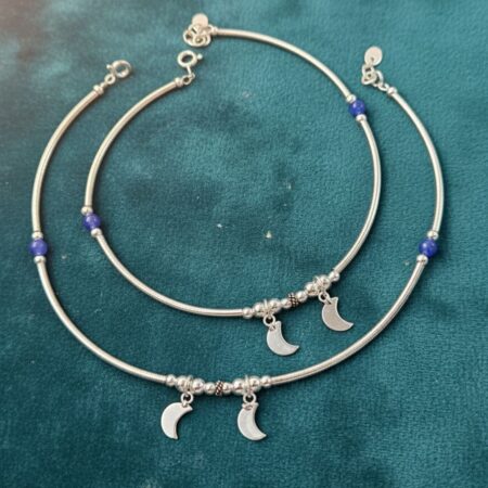 A studio shot of a pair of silver snake chain anklets (ankle bracelets) on a white background. Each anklet features a smooth, flexible silver chain, a single vibrant blue bead, and two small, dangling crescent moon charms next to a cluster of tiny silver beads. The anklets have an adjustable extension chain and lobster clasp closure.